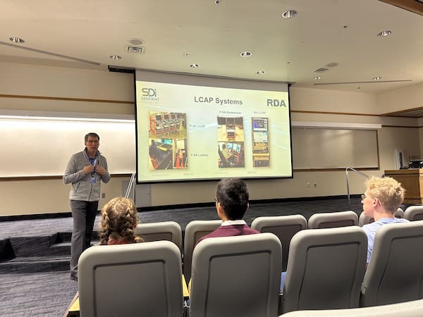 Jon Dionne stands beside a screen showing the LCAP system to an audience of students.